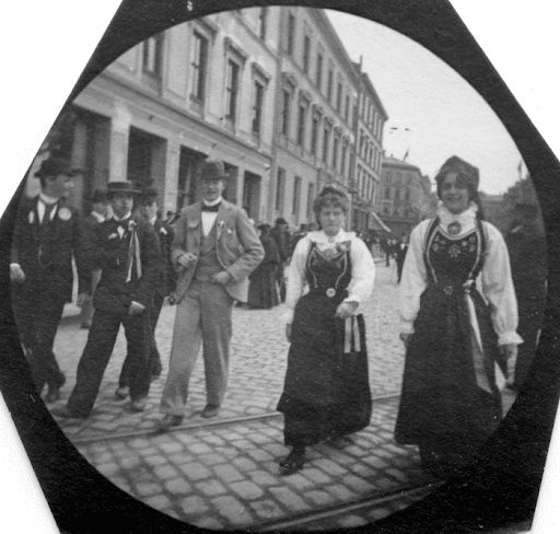 Mennesker på gaden, inklusive to kvinder i traditionelt norsk påklædning. En af dem smiler til kameraet