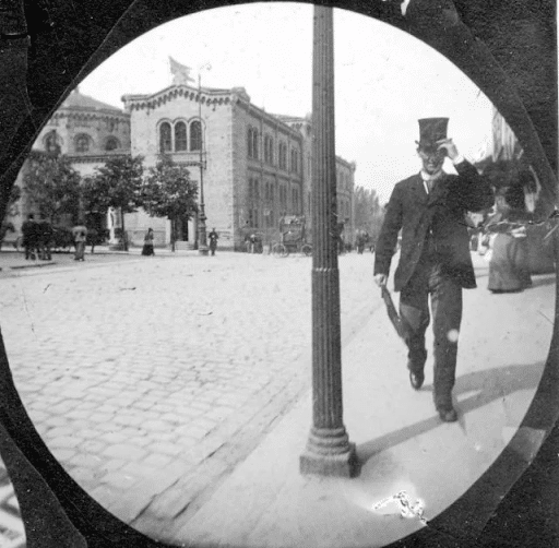 A man tips his hat as he passes a lamppost on the sidewalk of a cobblestone street. Across the road are some trees, an elegant building, and other people milling around.