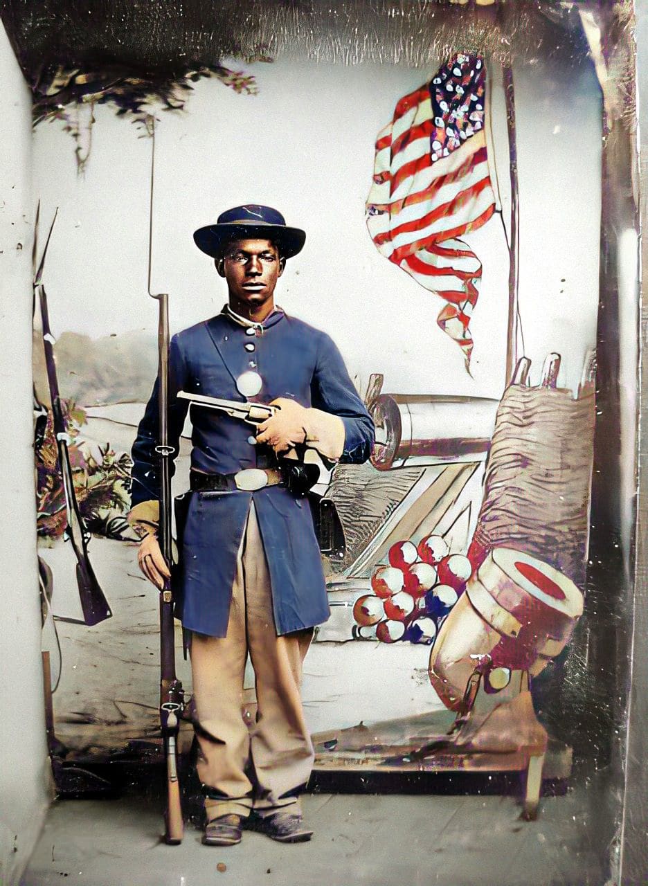 Unidentified Black soldier in Union uniform with a rifle and revolver in front of painted backdrop showing weapons and an American flag, sometime between 1863 and 1865. Courtesy of the Library of Congress